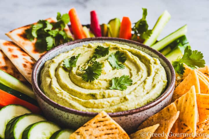 A smooth, green avocado dip in a bowl, topped with cilantro. The bowl is placed in the center of a platter, surrounded by vegetable crudités and pita chips.