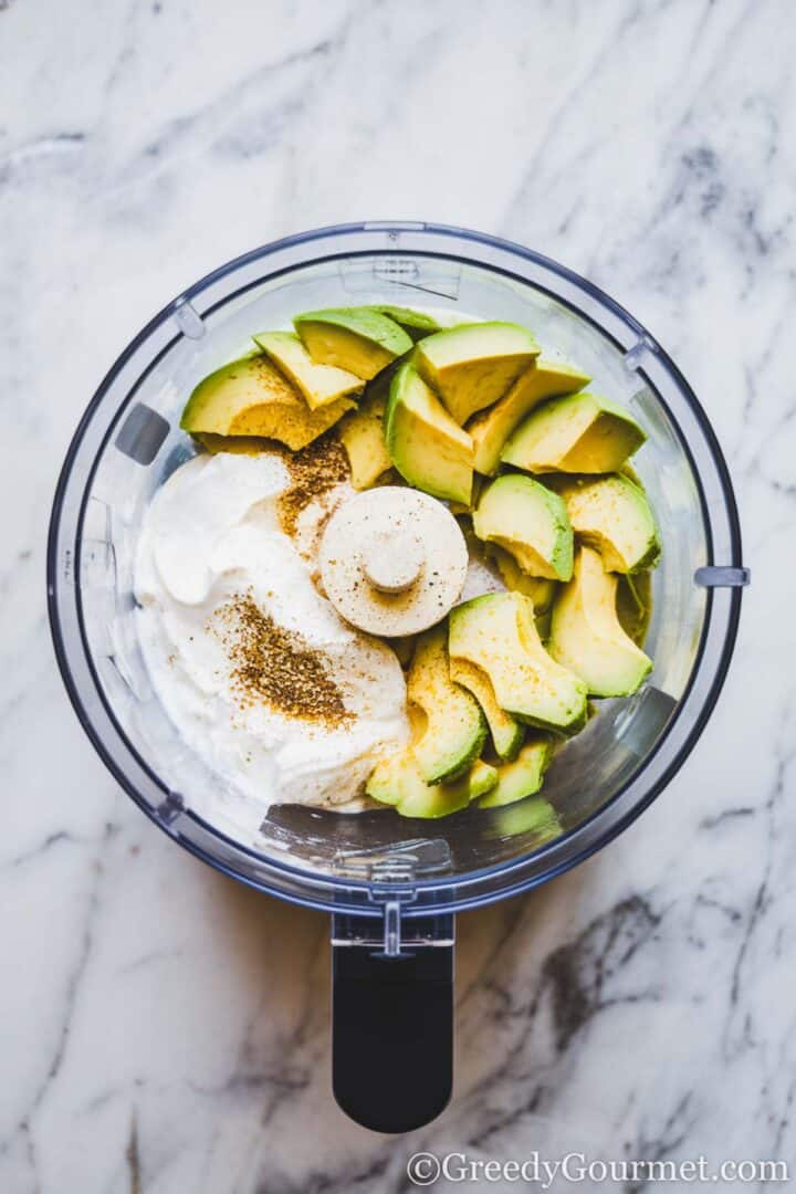 Processor bowl filled with cream cheese, avocado slices and seasoning, placed on a light grey marble surface.