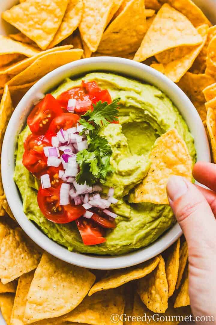 Bowl of smooth, green, avocado dip, topped with sliced cherry tomatoes, chopped red onions, cilantro. The bowl is surrounded by tortilla chips and tortilla chip is being dipped into green dip.