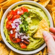 a bowl of creamy avocado dip, topped with chopped tomatoes, onions and cilantro, surrounded by tortilla chips