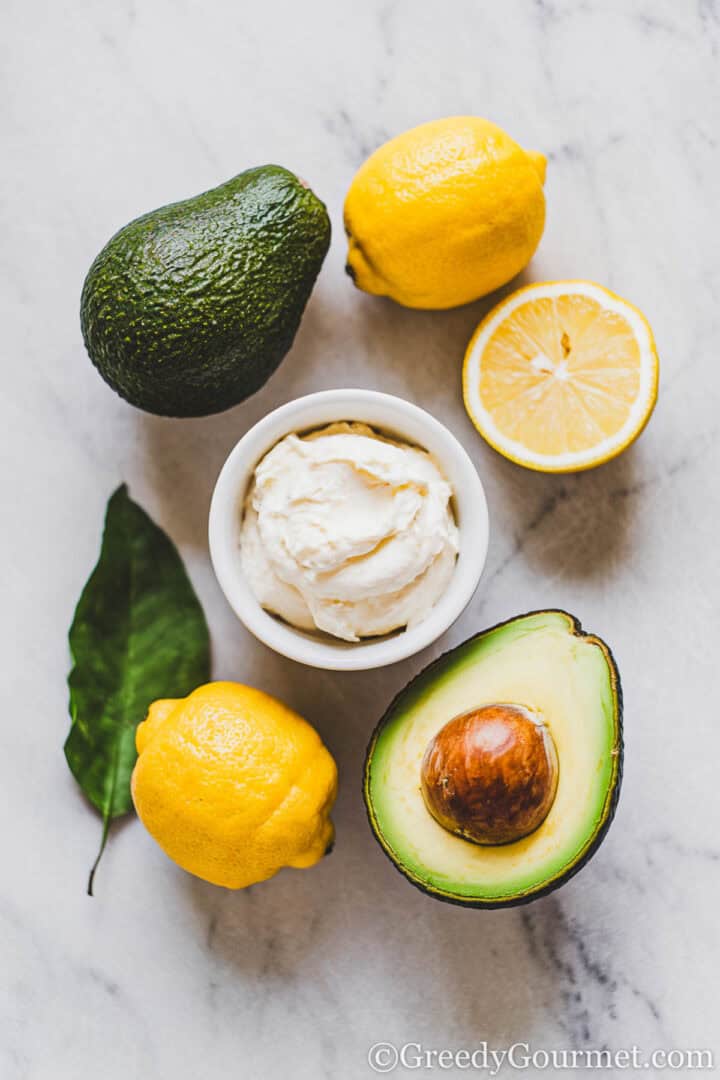 One whole avocado, one halved avocado, 2 lemons, half a lemon, green leaf, bowl of cream cheese. All of these items are placed on a light grey marble surface.