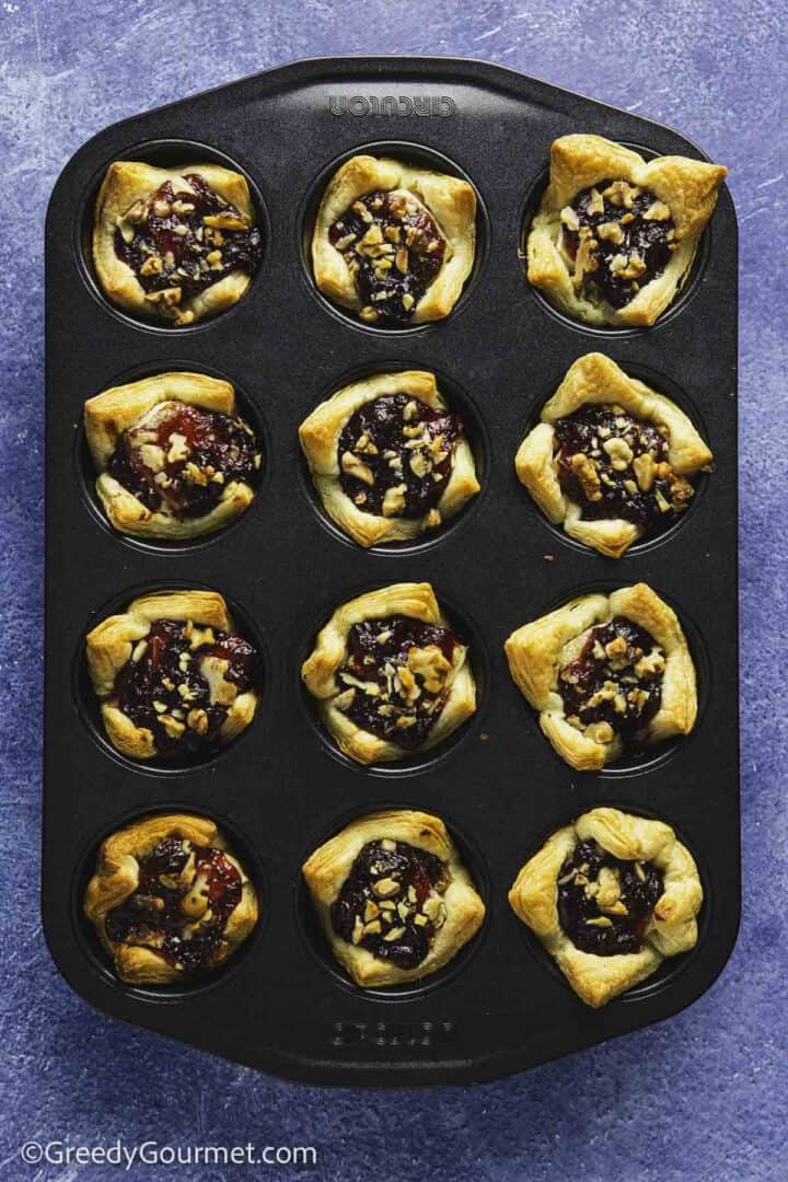 golden baked pastry cups with a jam filling and sprinkled nuts in a muffin tin on blue background