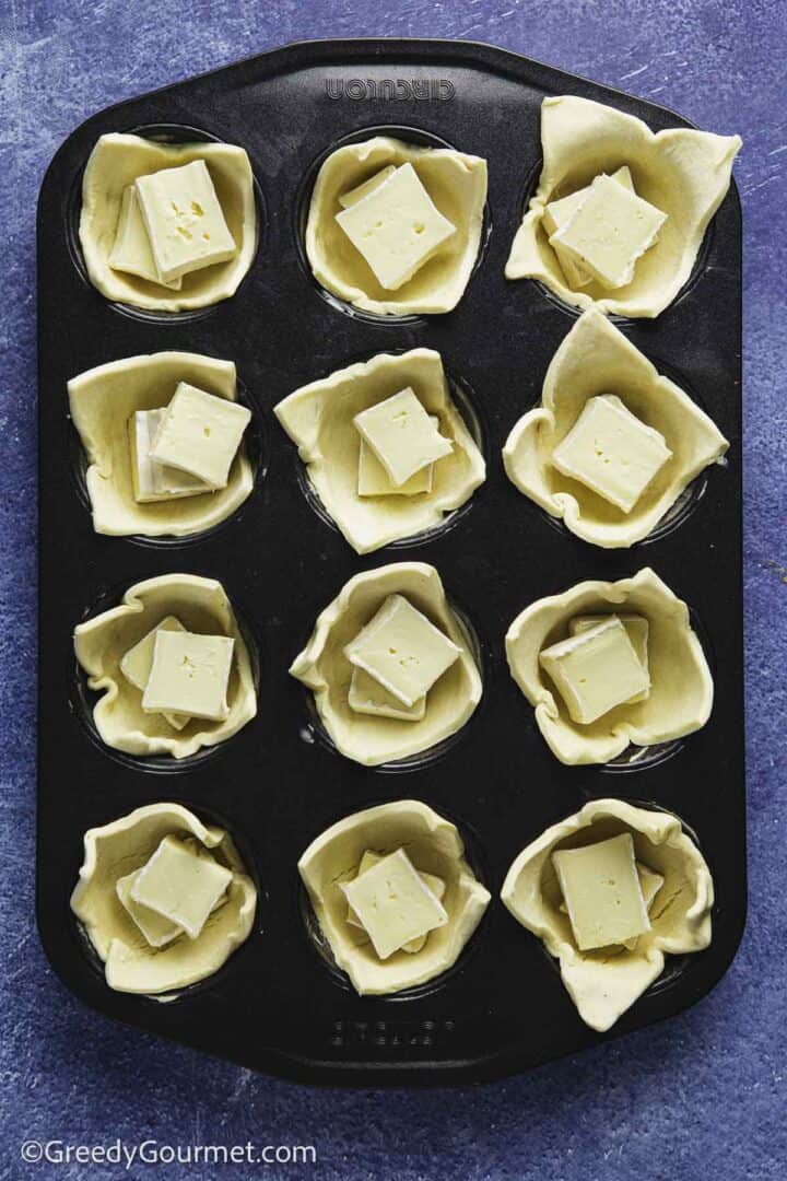 puff pastry cups in a muffin tin filled with wedges of cheese on blue table top