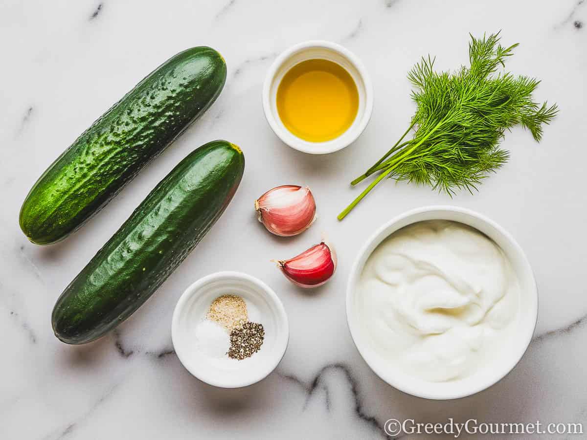 Cucumbers, garlic cloves, a small bowl of olive oil, another small bowl of salt and pepper, and a medium bowl of greek yogurt, arranged on a white marble top.