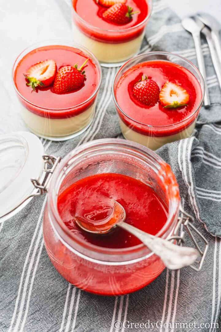 A half full glass jar of strawberry coulis with a spoon inside.