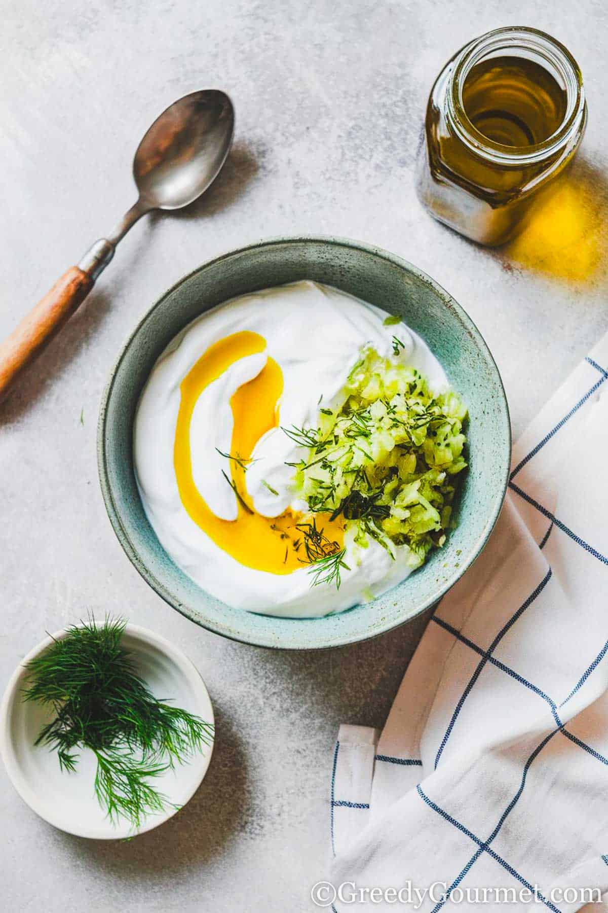 bowl filled with greek yogurt, grated cucumber, dill and olive oil. blue striped cloth, spoon, bowl of dill sprigs, and bottle of olive oil on the side