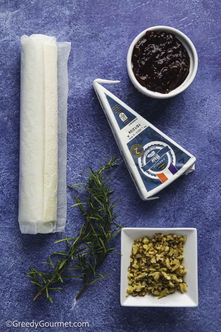 roll of puff pastry, wedge of brie, a bowl of cranberry sauce, a bowl of chopped walnuts, springs of rosemary, arranged on blue table top