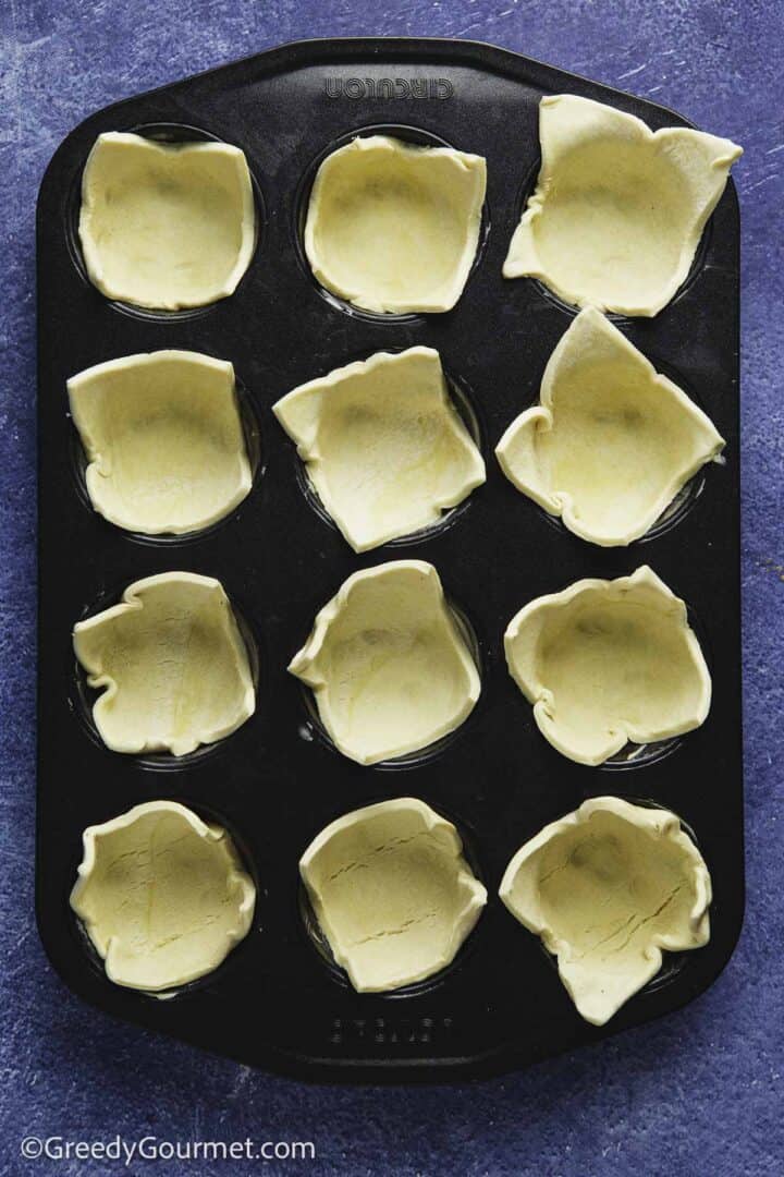 muffin tin filled with raw puff pastry squares on blue table top.