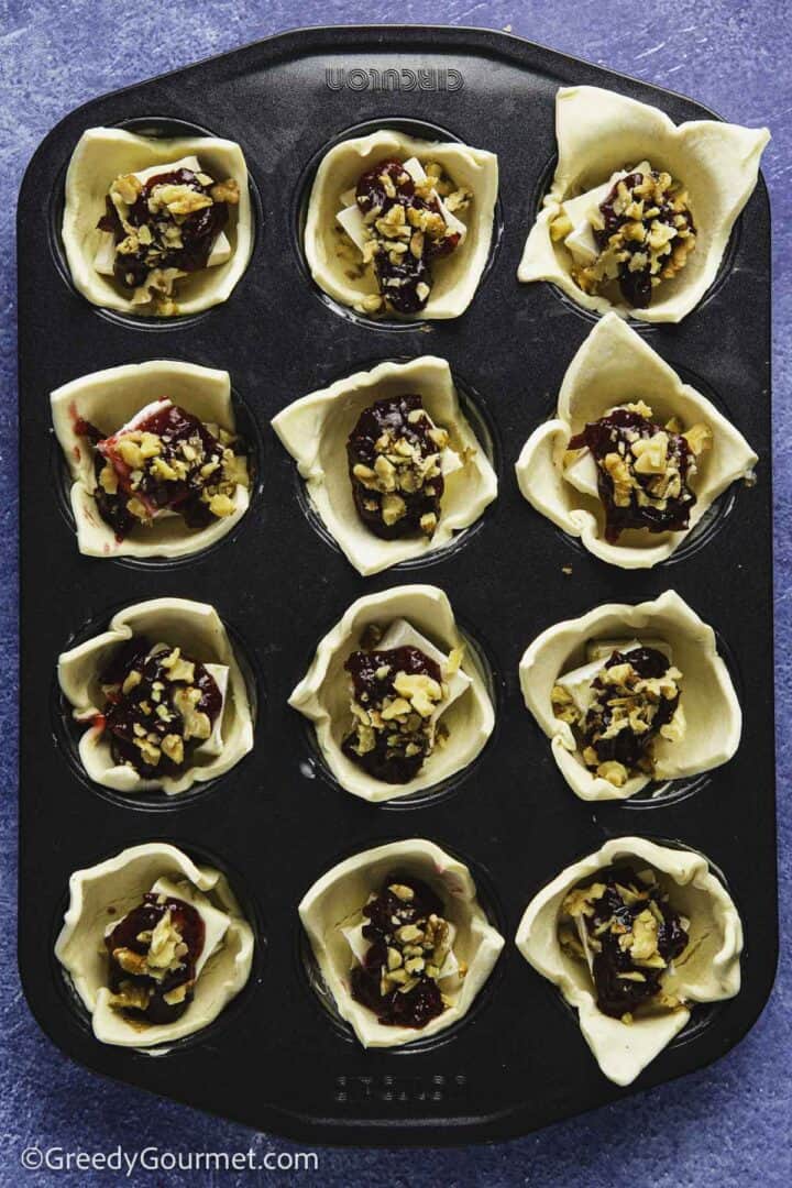 sprinkled nuts on jam in raw pastry cups in muffin tin on blue table top