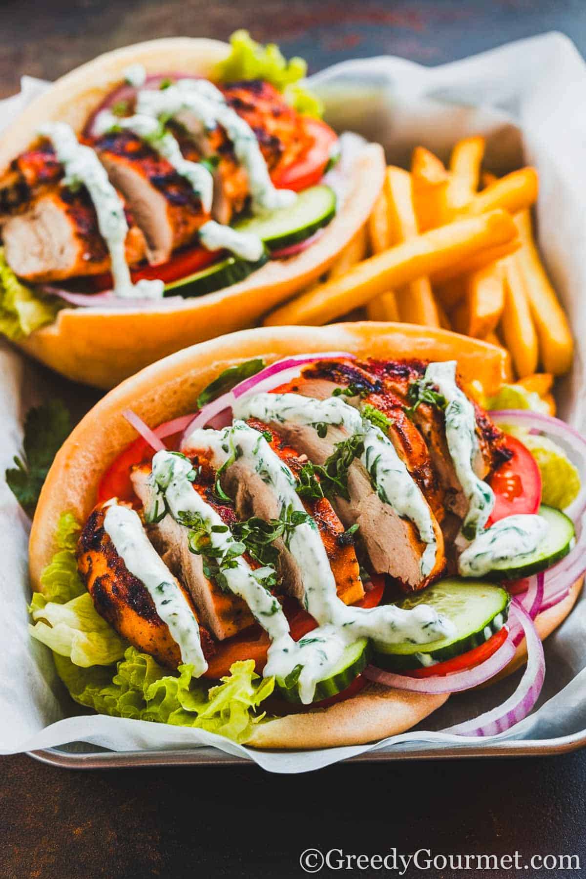 grilled, sliced chicken breasts inside a pita bread, along with cucumber, tomato, red onion, lettuce, drizzled with tzatziki. it's on a baking tray with fries.