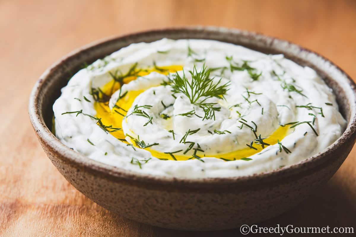 a bowl filled with creamy tzatziki sauce, drizzled with olive oil and finely chopped dill on a wooden table